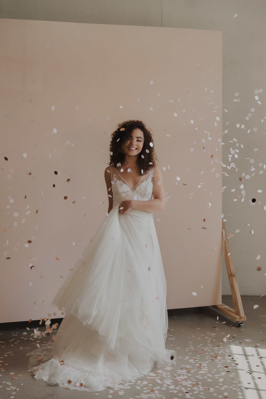 black-curly-hair-bride-wearing-white-sleeveless-dress-with-confetti-in-studio-pink-wooden-backdrop