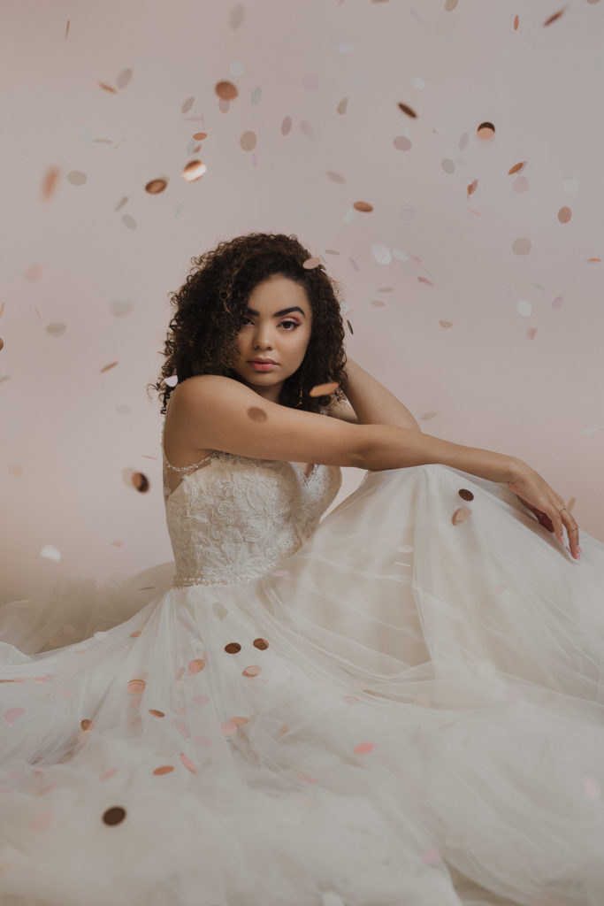 bride-sitting-down-in-blush-wedding-gown-with-pink-confetti-in-studio-curly-hair-black
