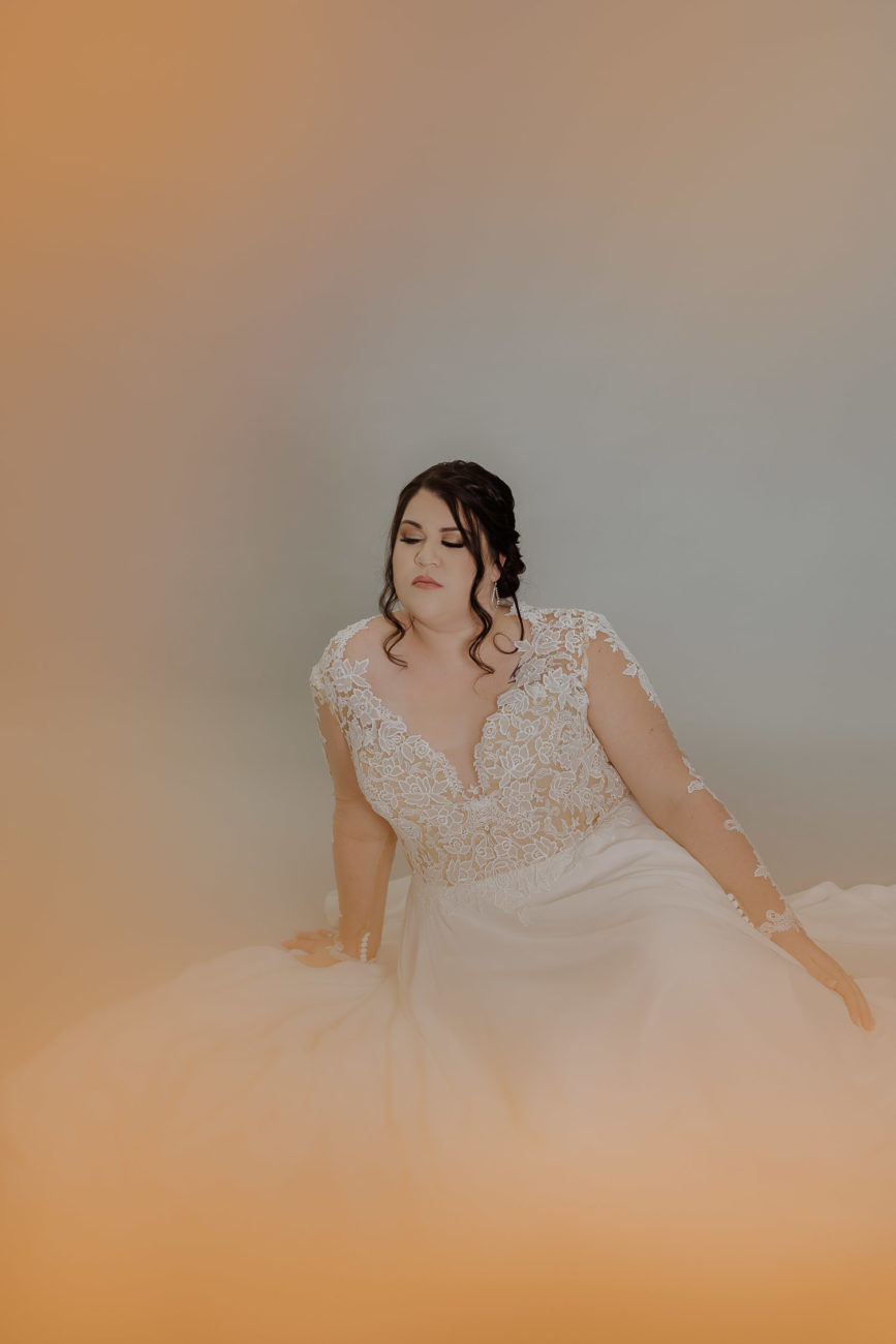 bride-wearing-long-sleeve-lace-wedding-dress-plus-size-updo-against-grey-peach-background